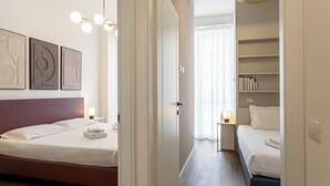 A hotel room with a bed, bedside tables, a bookshelf, and a hallway leading to another room.