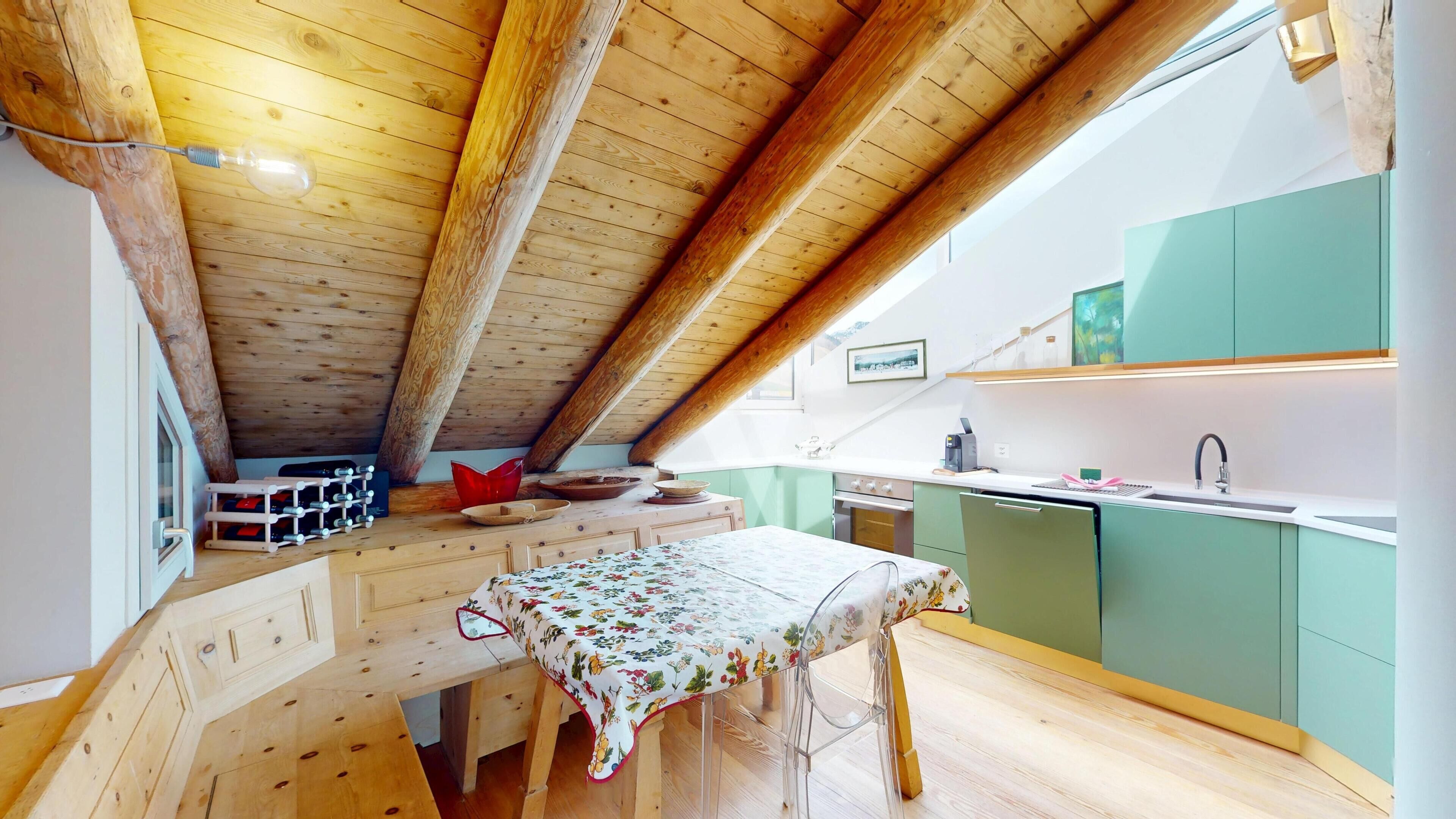 A cozy attic room with a wooden ceiling, a dining table with a floral tablecloth, and a kitchen area with green cabinets and a window.