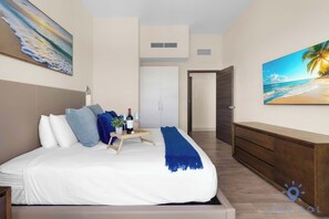 A bedroom with a bed, a bedside table with a wine bottle and glasses, a blue throw blanket, and a painting of a beach scene.