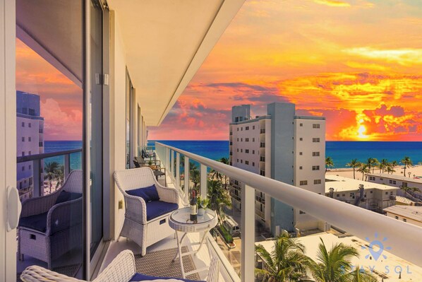 A balcony with wicker furniture and a small table, overlooking a beach and cityscape during sunset.