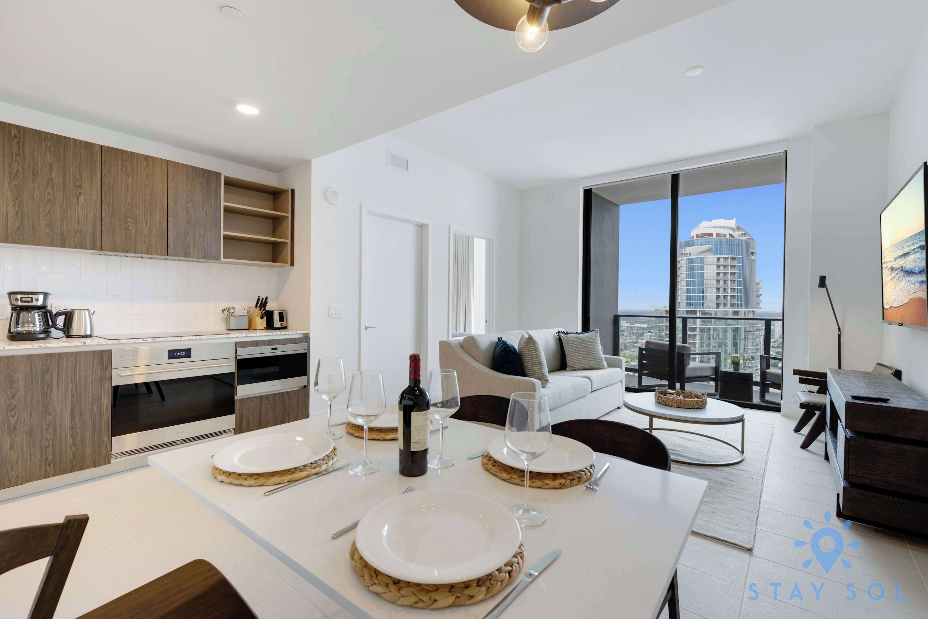 Una cocina moderna con armarios de madera, un comedor preparado para cuatro personas con copas de vino y una botella, y una sala de estar con sofá y vistas a un paisaje urbano.
