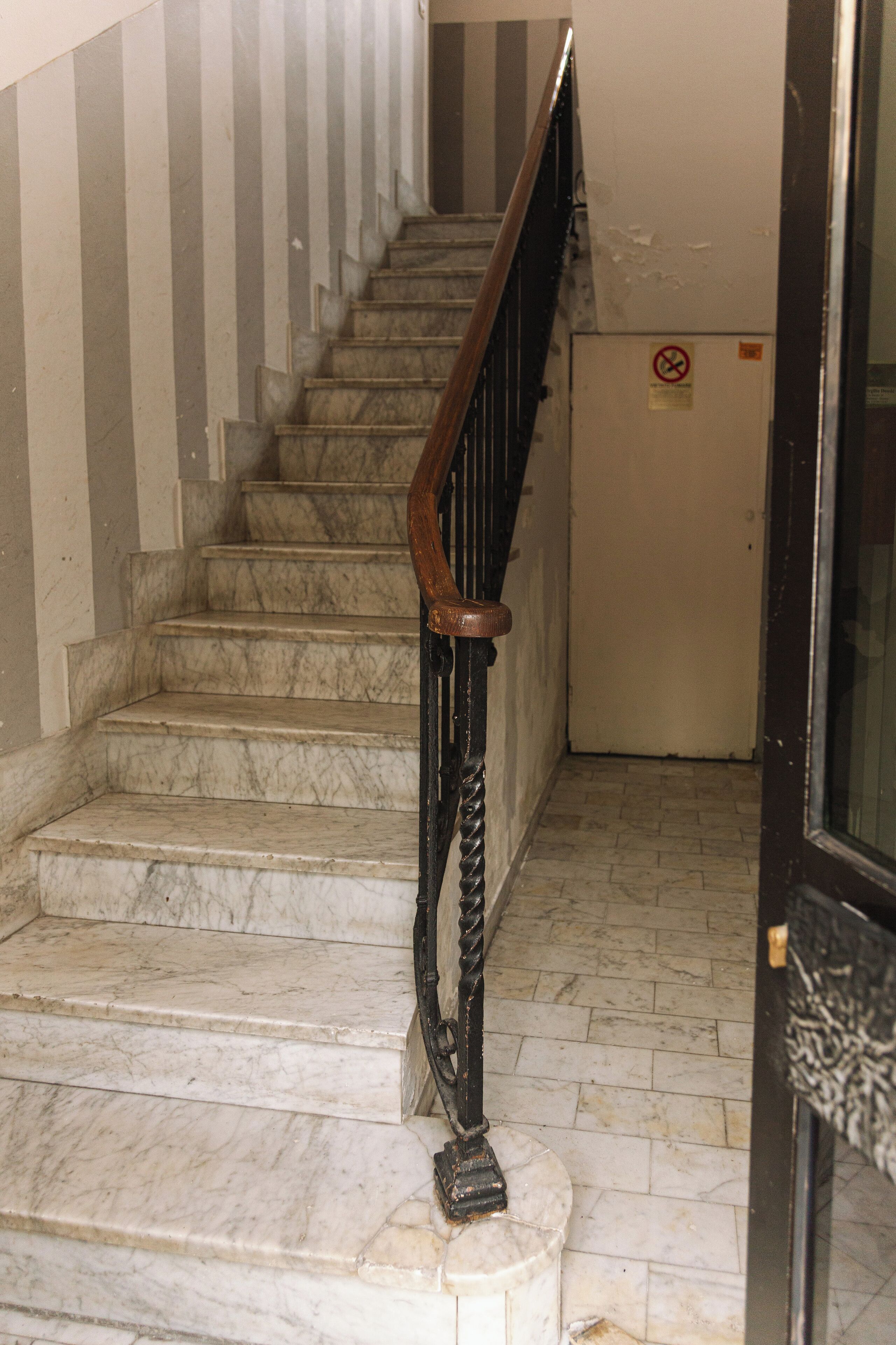 Una scala in marmo con ringhiera nera che conduce a una porta con il cartello 'no smoking'.