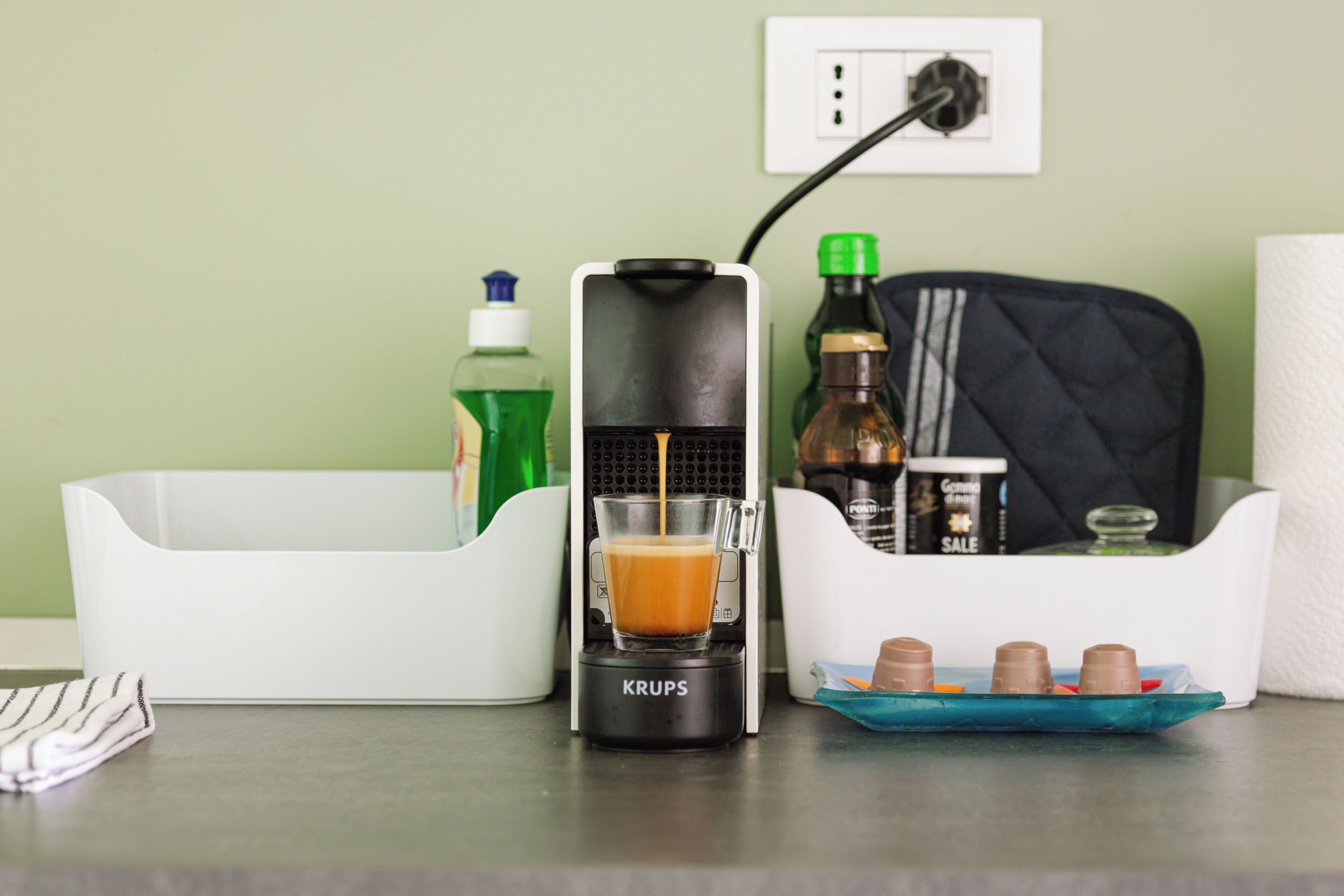 A coffee maker with a glass of coffee, a tissue roll, and a tray with chocolate cups on a table.