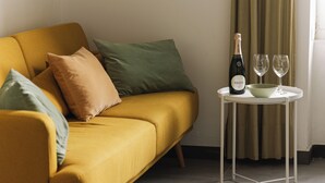 A mustard-colored sofa with multiple cushions, a small white table with a bottle and two glasses, and a green bowl.