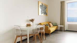 A modern living room with a white dining table, clear chairs, a yellow sofa, and a view of the sea through a large window.