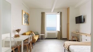 A hotel room with a bed, a desk with a computer, a sofa, a dining table with chairs, a view of the sea, and a painting on the wall.