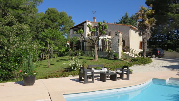 A poolside area with outdoor furniture, a swimming pool, and a house with a stone facade and a staircase leading to the entrance.