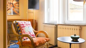 Een kamer met een houten stoel met een kleurrijk kussen, een zwarte vloerlamp, een klein rond tafeltje met een groen voorwerp, en een raam met uitzicht op een gebouw.