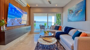 Una sala de estar moderna con un televisor grande, un ventilador de techo, un sofá con cojines coloridos y un cuadro de tortugas marinas.