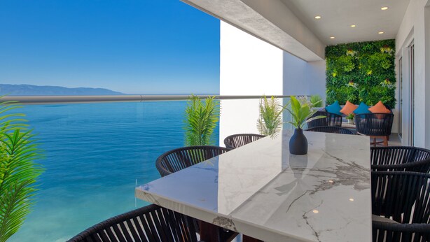 Un balcón con vista al mar, una mesa de mármol, sillas de ratán negro y una pared verde con plantas.