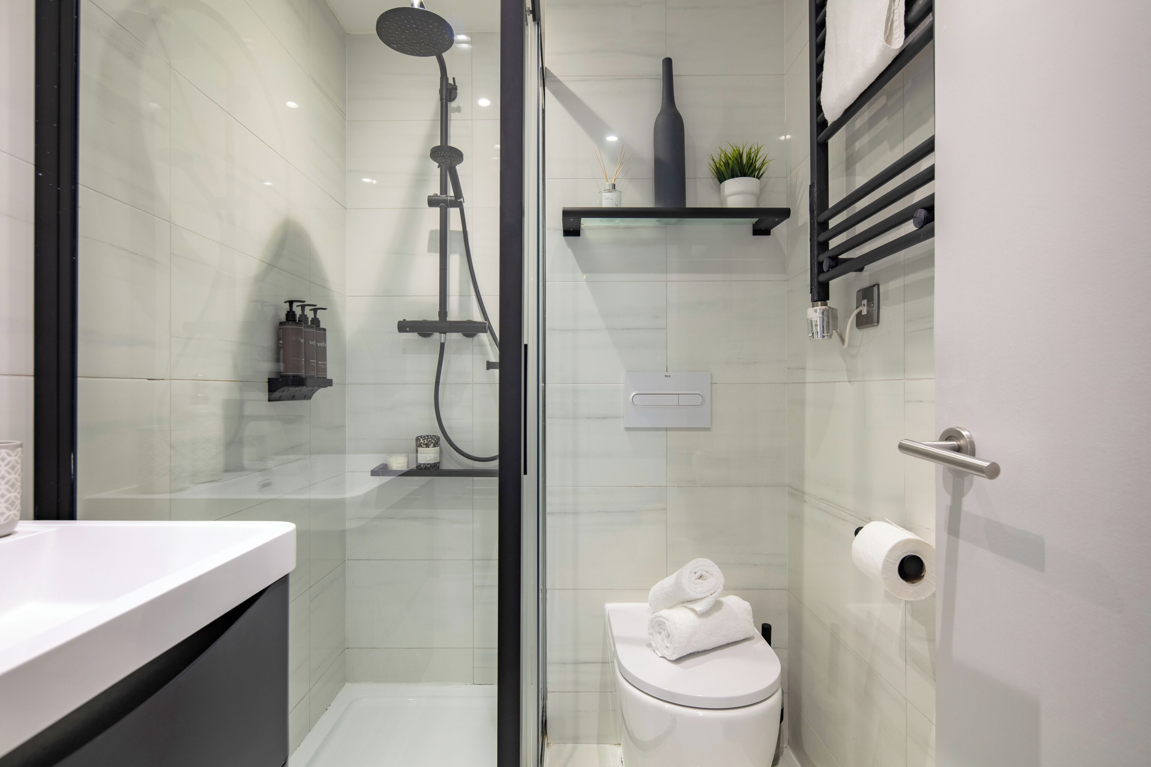 A modern bathroom with a glass-enclosed shower, a toilet with a towel on top, and a shelf with toiletries.