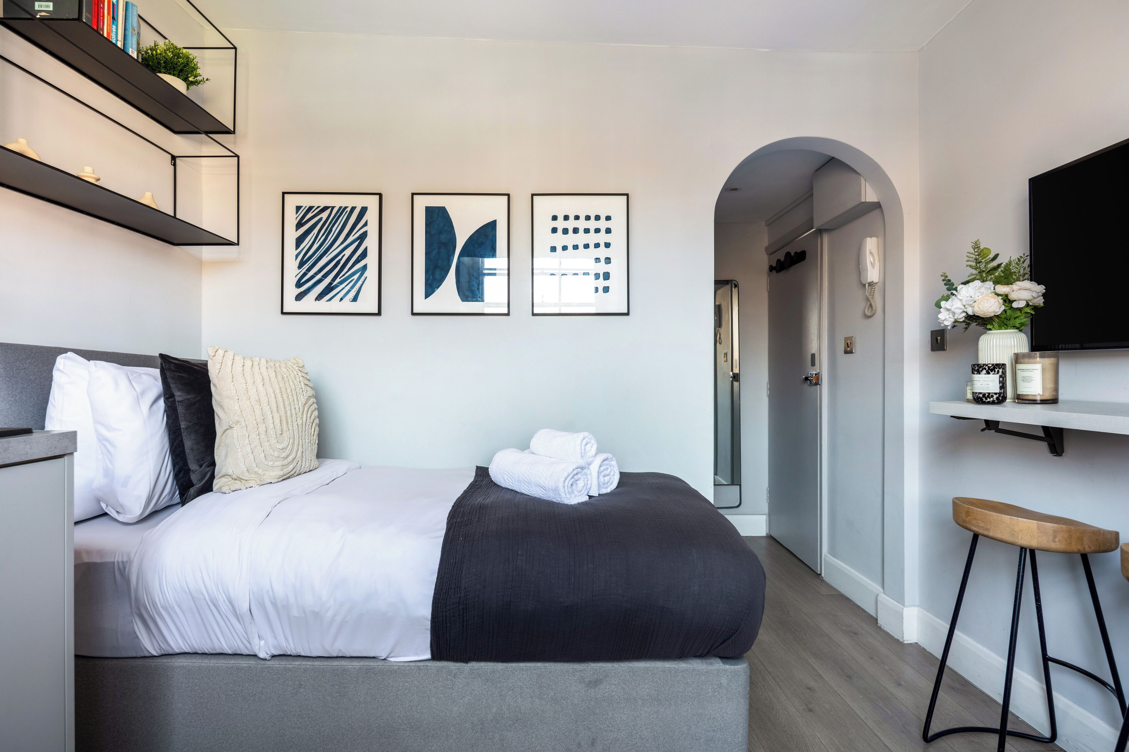 A modern hotel room with a bed, a TV, a desk, and a bathroom visible through an archway.