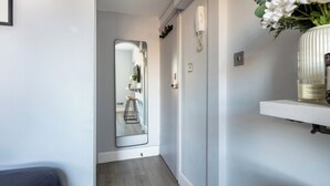 Un corridoio con uno specchio che riflette una stanza, una porta bianca e uno scaffale con un vaso di fiori.