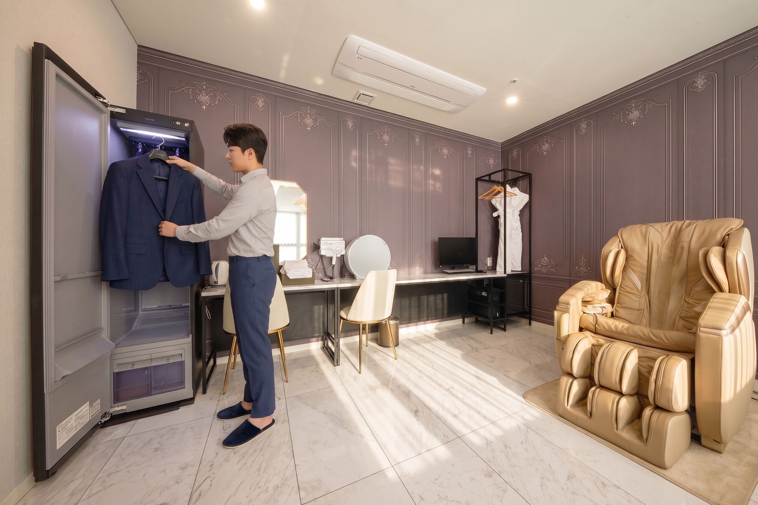 A man in a room with a built-in wardrobe, a desk with a computer, a massage chair, and a vanity with a mirror.
