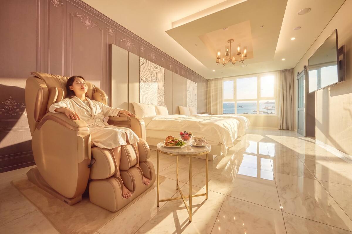A room with a massage chair, a bed, a small table with food, and a large window with a view.