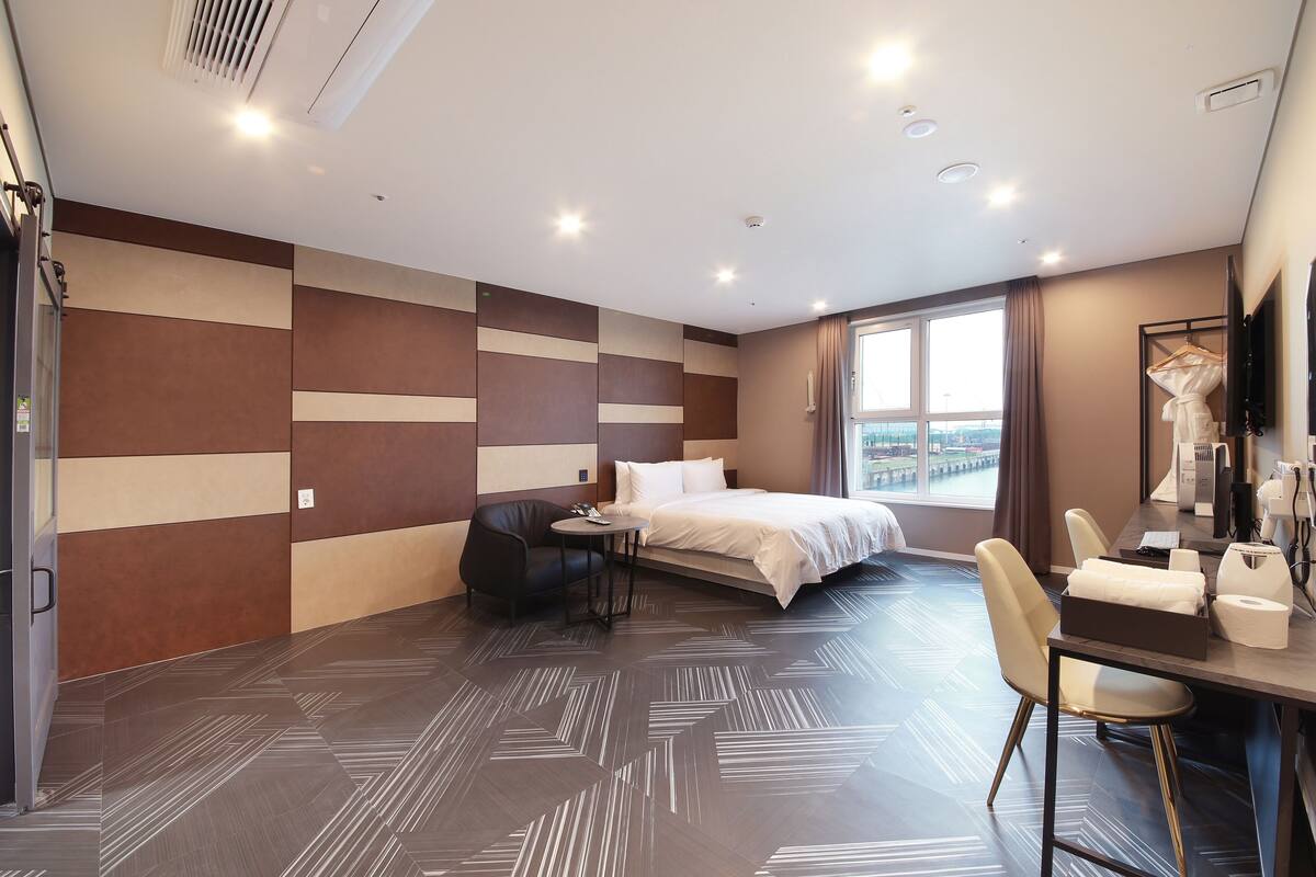 A modern hotel room with a bed, desk, and chair, a large window with a view, and a mirror.