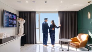 A hotel room with a large window, a flat-screen TV, a robe on a rack, a small round table, and a leather armchair. - Restia Incheonbay Hotel (Incheon)