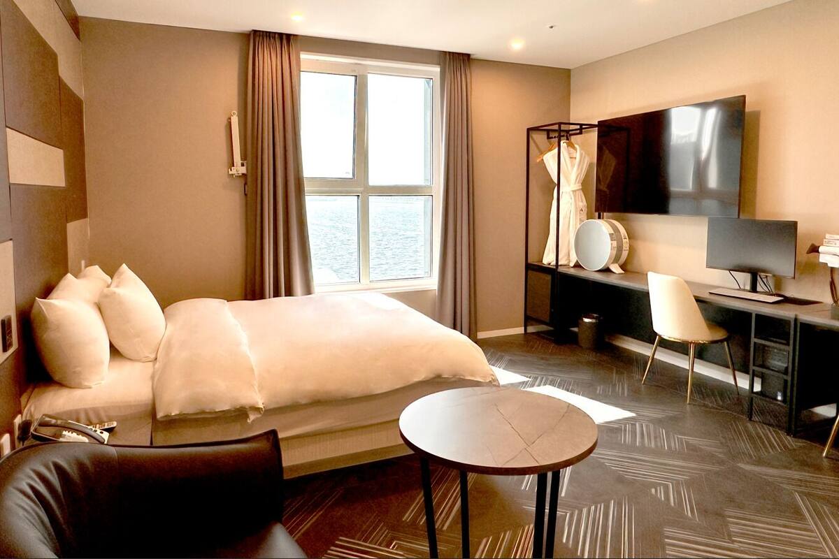 A hotel room with a large bed, a desk with a chair, a TV, and a view of the water through the window.