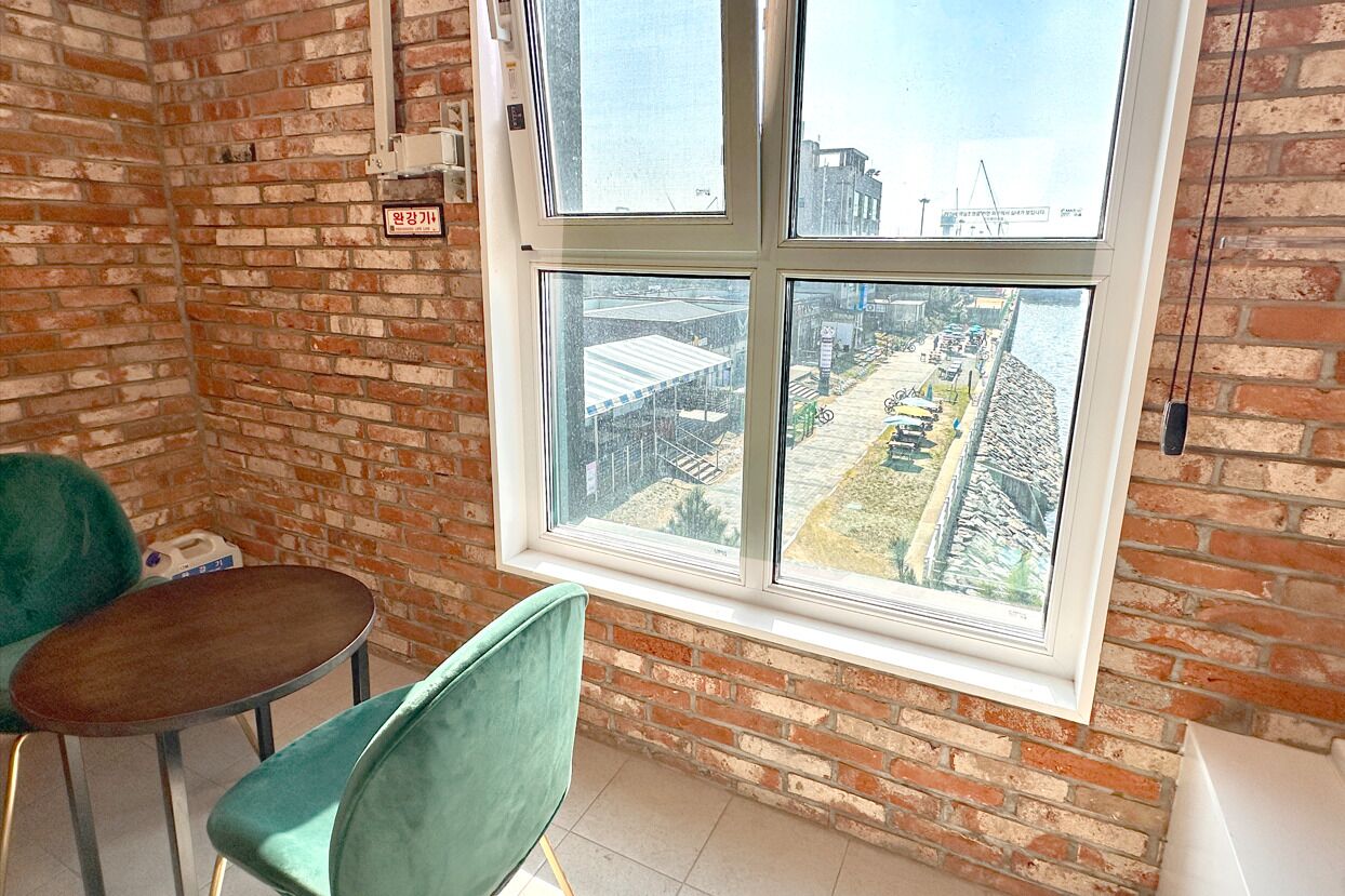 A room with a brick wall, a window with a view of a construction site, a green chair, and a small table.