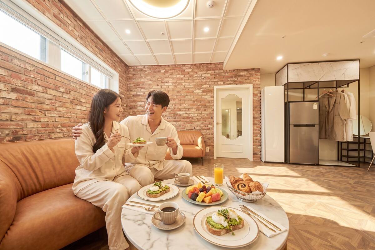 A modern room with a brick wall, a sofa, a dining table with breakfast, a refrigerator, and a bathroom with a mirror and towel rack.