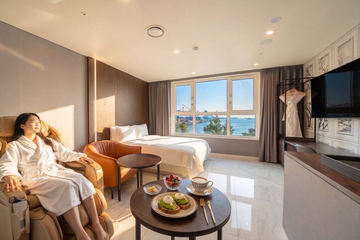 A hotel room with a massage chair, a bed, a dining table with breakfast, and a large window with a view of the sea.