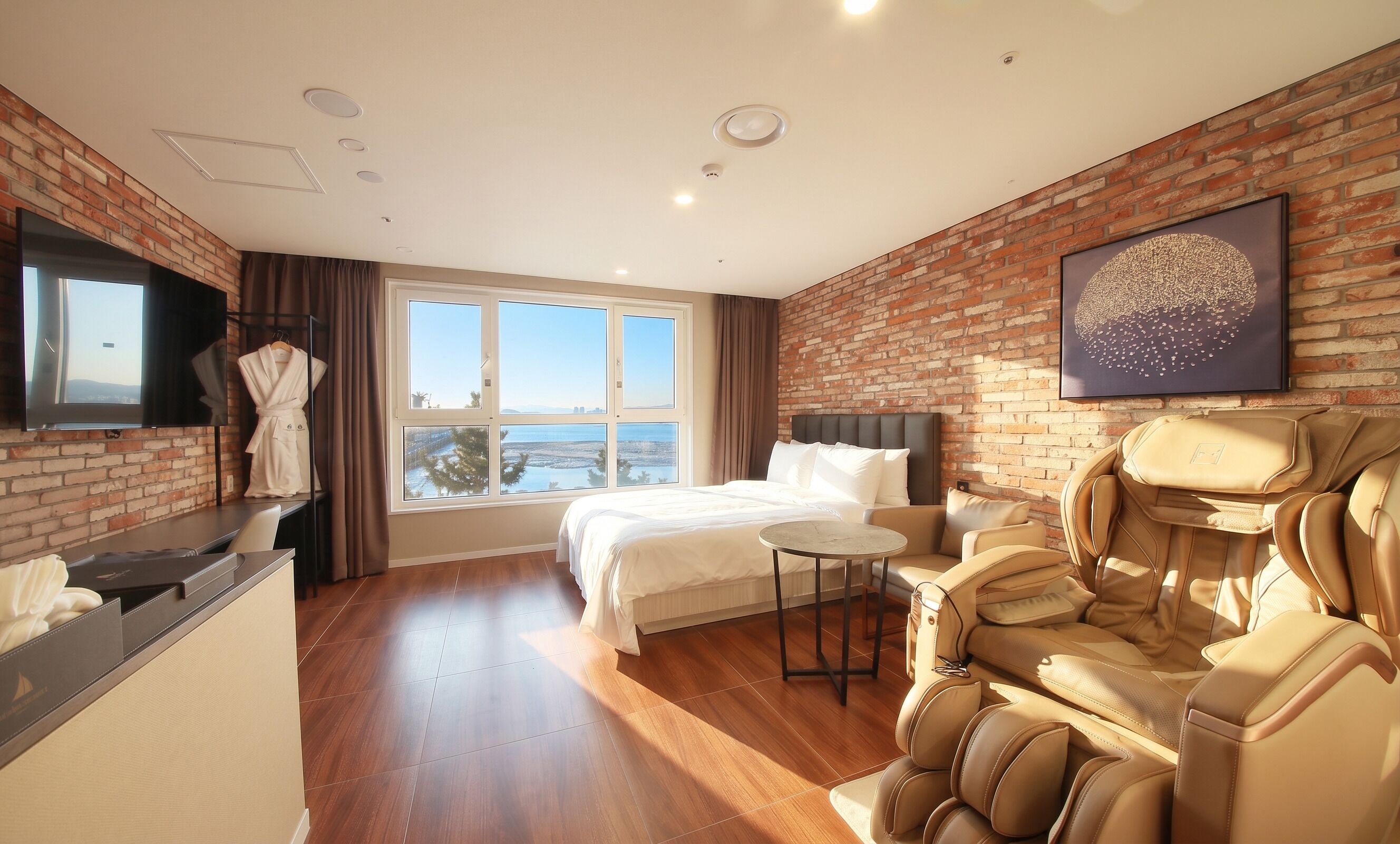 A hotel room with a bed, massage chair, large window, and brick wall.