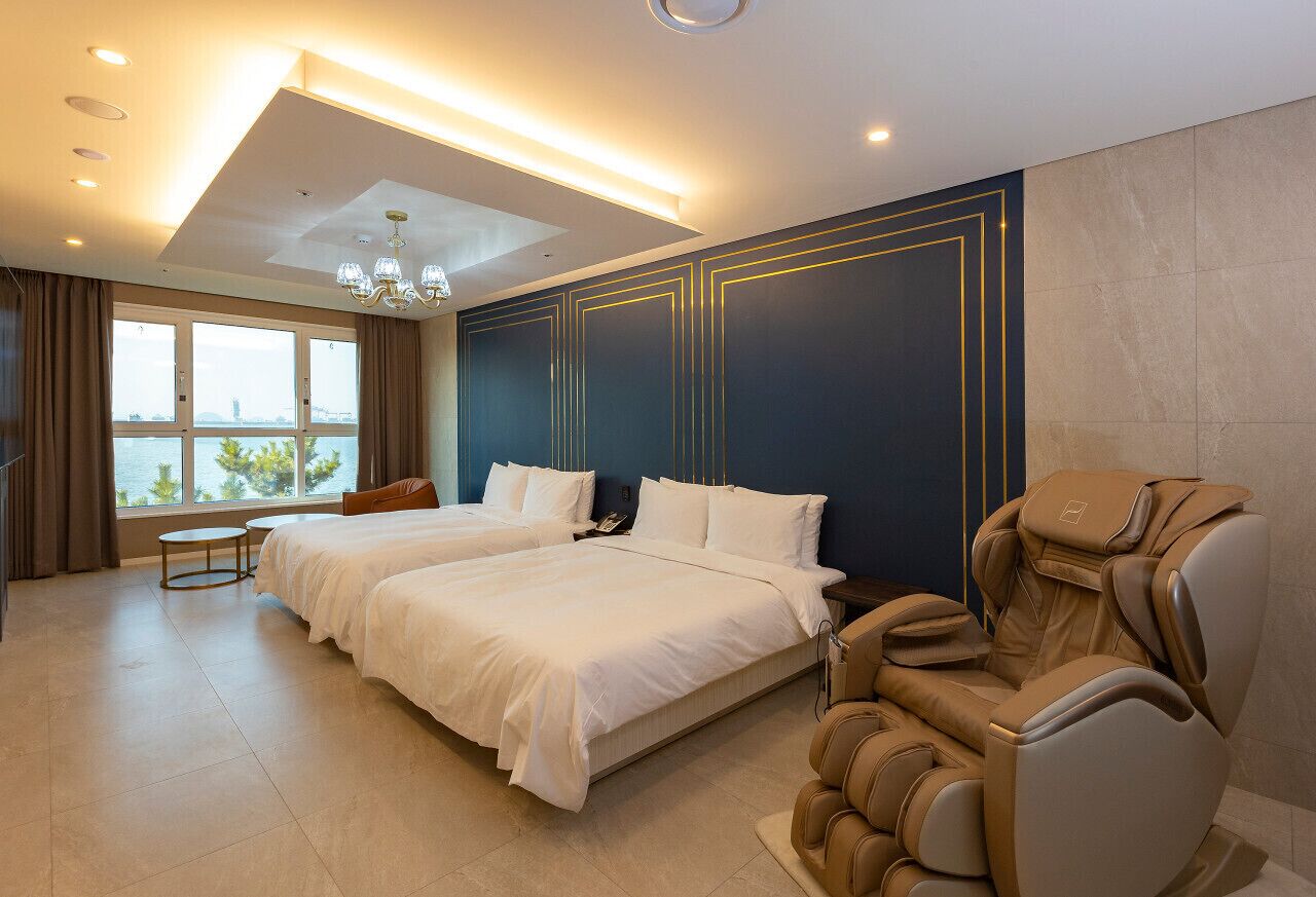 A hotel room with two beds, a massage chair, a small table, and a view of the city.