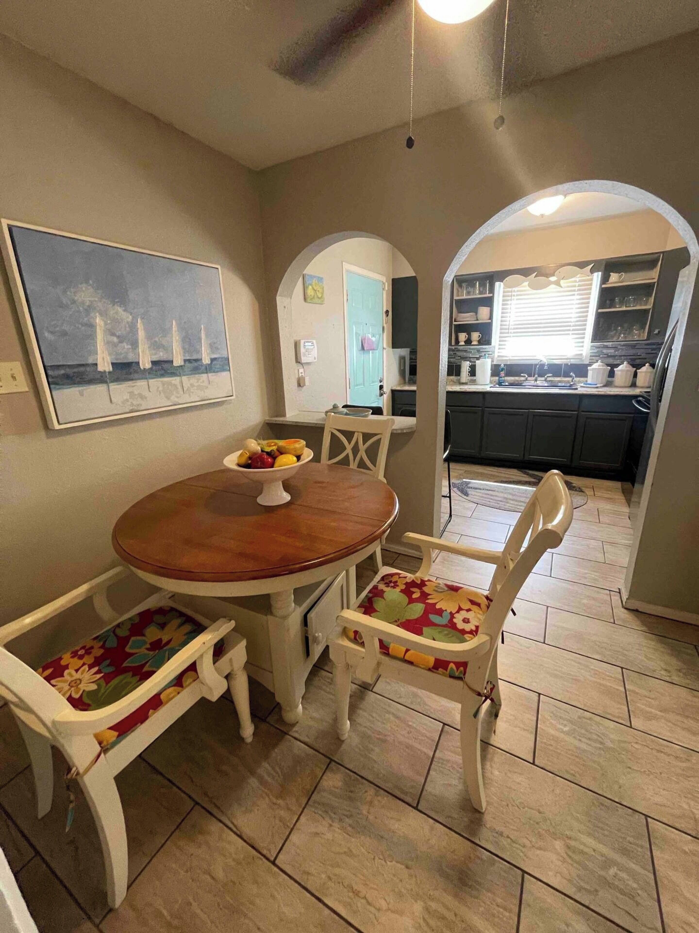 Zona pranzo con tavolo in legno e sedie bianche, un quadro appeso alla parete e una cucina sullo sfondo.