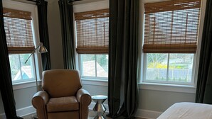 A hotel room with a bed, a chair, a small table, and two windows with blinds.
