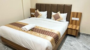 A neatly made bed with a brown headboard, white bedding, and patterned pillows. There are two bedside tables with wooden sides and a power outlet on each. The room has a marble floor. - HOTEL SHREE HARI (Dwarka)