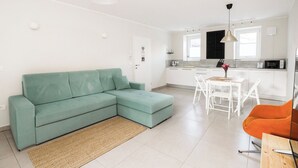 Ein modernes Wohnzimmer mit einem Ecksofa, einem Esstisch mit Stühlen, einer Küchenzeile inklusive Mikrowelle und einem kleinen orangefarbenen Sessel.