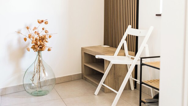 Ein Zimmer mit einer Glaskaraffe voller getrockneter Zweige, einem weißen Stuhl, einem Holzregal und einer horizontalen Holzverkleidung.