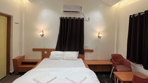 Habitació d'hotel amb un llit gran, un ventilador de sostre, un aparell d'aire condicionat, dues làmpades laterals, una tauleta de nit de fusta, una cadira vermella i una finestra amb cortines.
