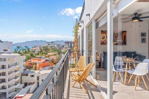 A balcony with wooden chairs, a dining table with chairs, and a view of the cityscape and mountains.
