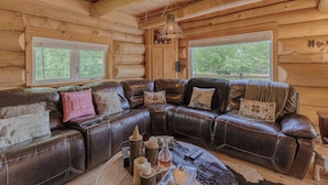 Ein gemütliches Blockhaus-Interieur mit einem großen, ledernen L-Sofa, zwei Fenstern und einer hängenden Leuchte.