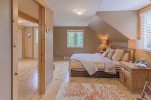 A bedroom with a large bed, wooden floor, a window with blinds, a bedside lamp, and a nightstand.