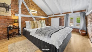 Un dormitorio con pared de ladrillo, techo inclinado, cama con colcha blanca y gris, mesita de noche con lámpara y ventana con cortinas.