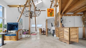 Amplia habitación con techo alto, vigas de madera a la vista, lámpara colgante, zona de comedor con mobiliario de mimbre y vistas a vegetación exterior.