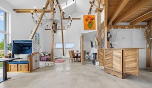 A spacious room with a high ceiling, exposed wooden beams, a hanging light fixture, a dining area with wicker furniture, and a view of greenery outside.