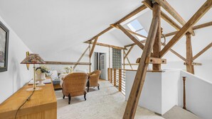 Una habitación con techos altos y vigas de madera a la vista, un escritorio con lámpara, sillas de mimbre y vistas a través de un tragaluz.
