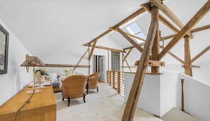 Ein Zimmer mit hohen Decken und freiliegenden Holzbalken, einem Schreibtisch mit Lampe, Korbstühlen und Blick durch ein Dachfenster.