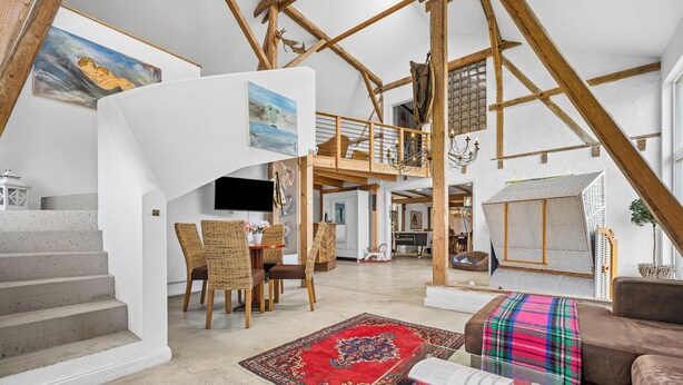 Amplio espacio tipo loft con vigas de madera expuestas, comedor con mobiliario de mimbre y zona de estar acogedora con una manta colorida.