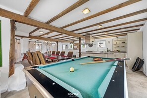 A pool table with a green felt surface, wooden beams on the ceiling, a dining area with chairs and a table, and a kitchen area with cabinets and a refrigerator.