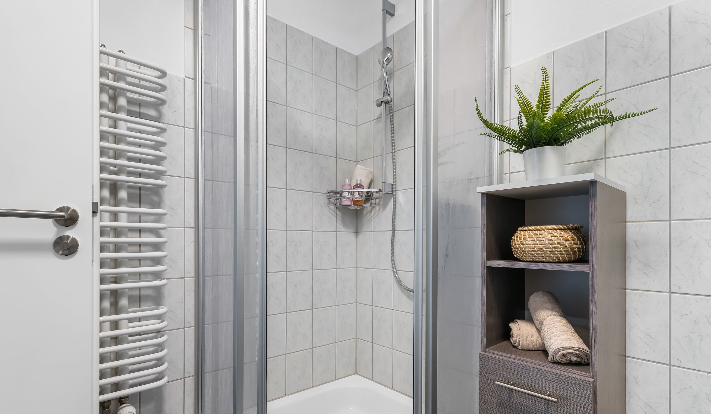 A modern bathroom with a glass shower enclosure, a towel rack, a shelf with towels, and a potted plant.