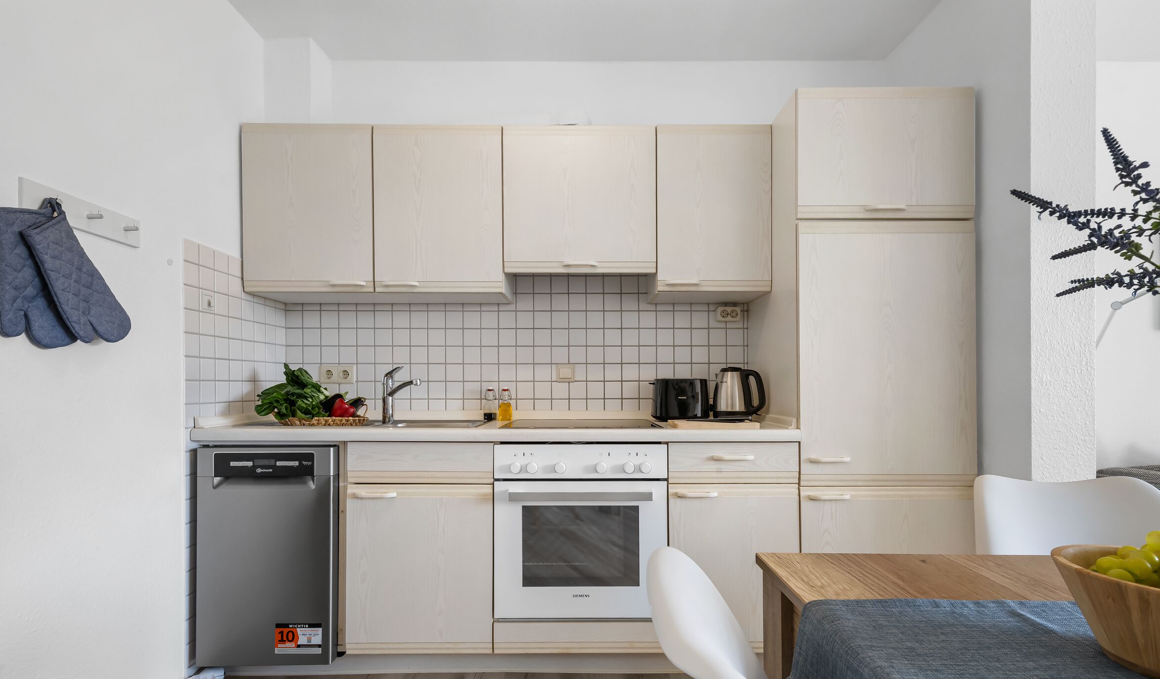 A compact kitchen with white cabinetry, a Siemens oven, a dishwasher, and a small dining area with a wooden table and a bowl of fruit.