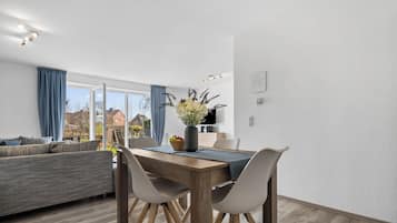 Ein modernes Wohnzimmer mit einer Couch, einem Esstisch und Stühlen. Ein großes Fenster mit Vorhängen, eine Vase mit Blumen und Blick auf Häuser außen.