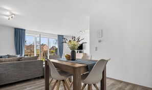 Una sala de estar moderna con un sofá, una mesa de comedor y sillas. Hay un ventanal con cortinas, un jarrón con flores y vistas a casas.