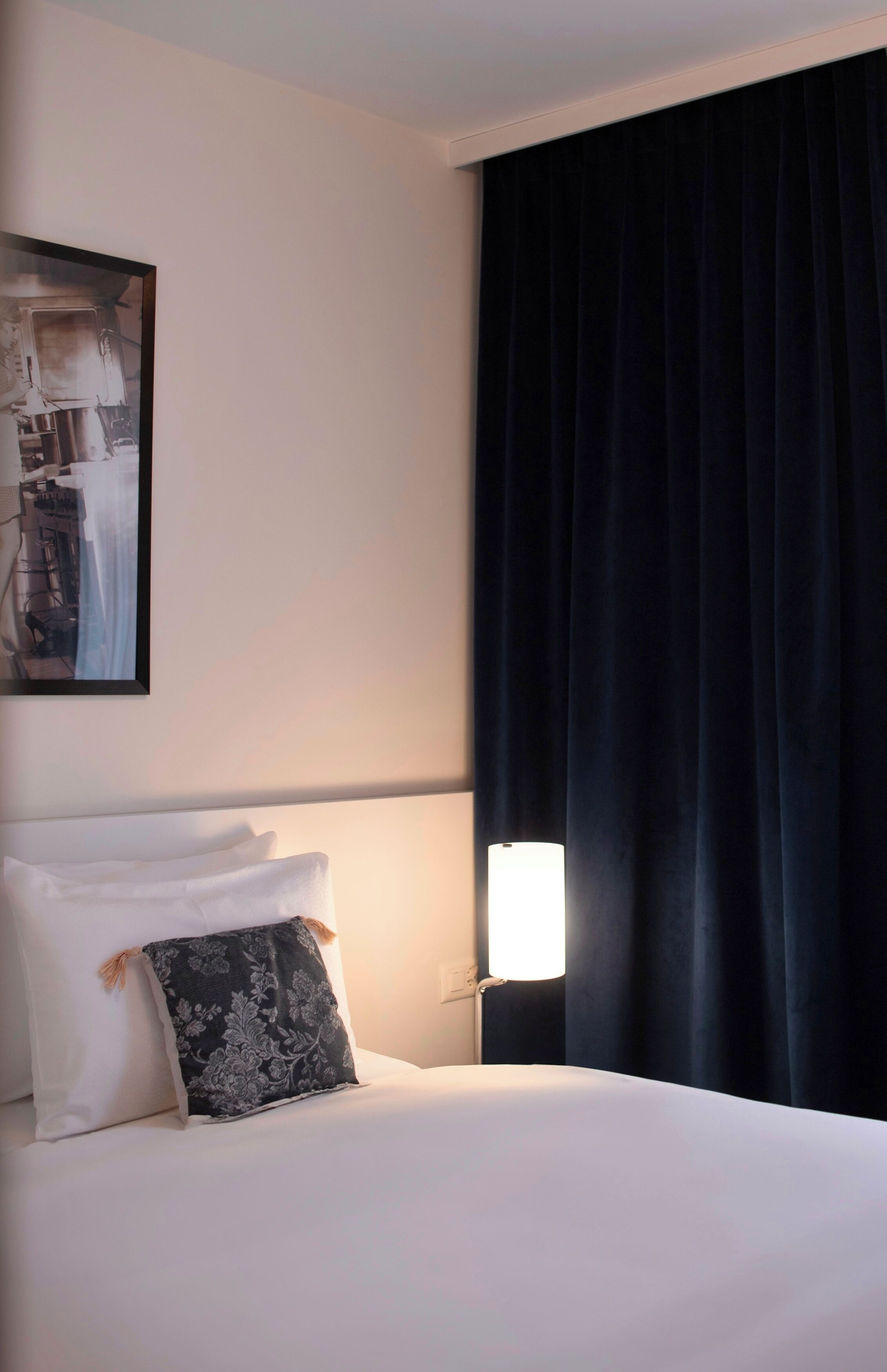 A hotel room with a neatly made bed, a framed picture on the wall, and a dark curtain covering a window.