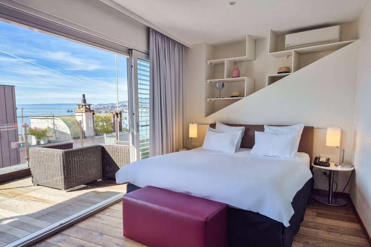 Un cuarto de hotel con una cama grande, un balcón con una silla de mimbre y vista al mar.