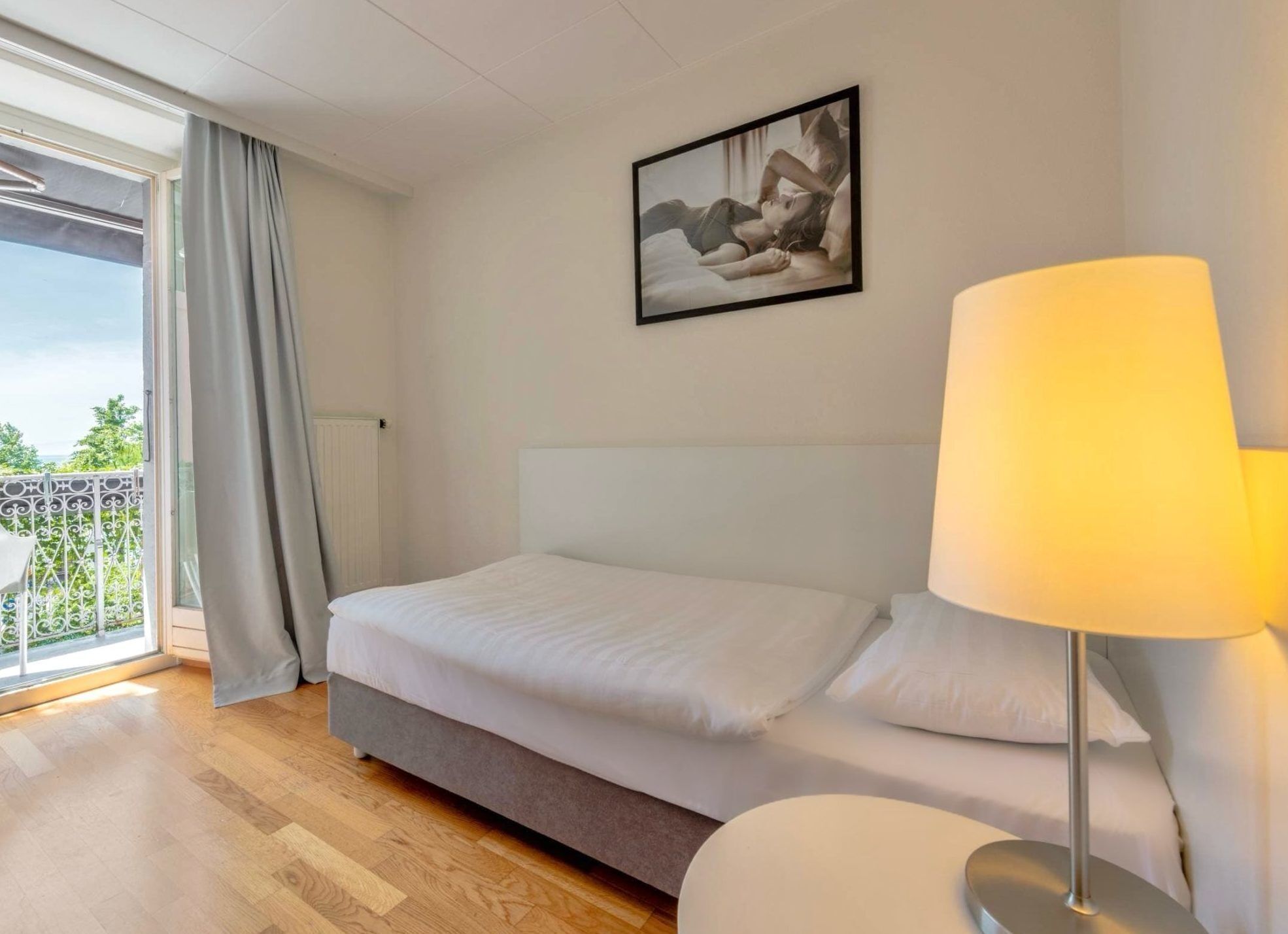 Un dormitorio con cama, lámpara de noche, un cuadro en la pared y vistas al exterior a través de una ventana.